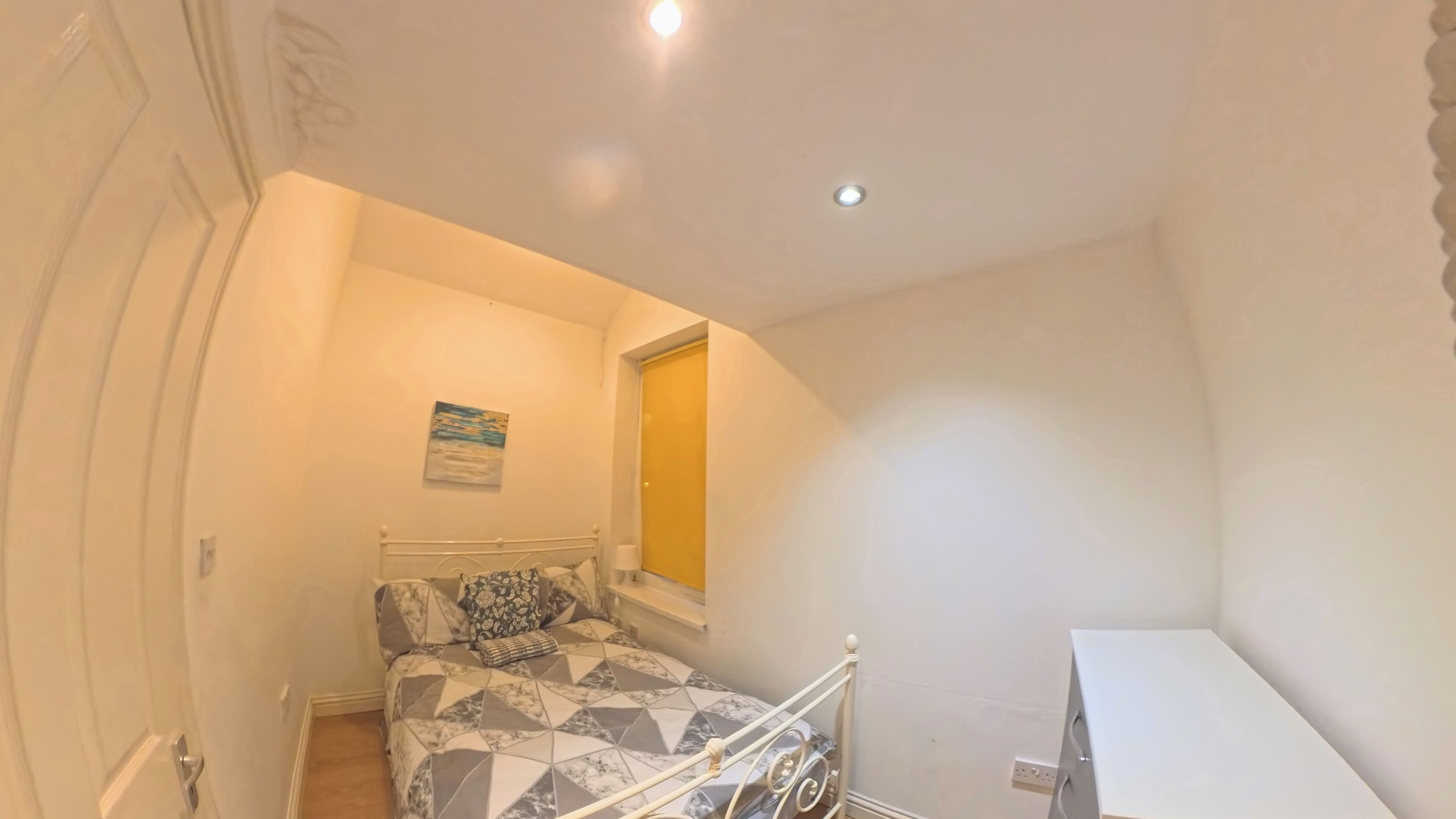A small, well-lit bedroom with a bed, a window, and a white desk.