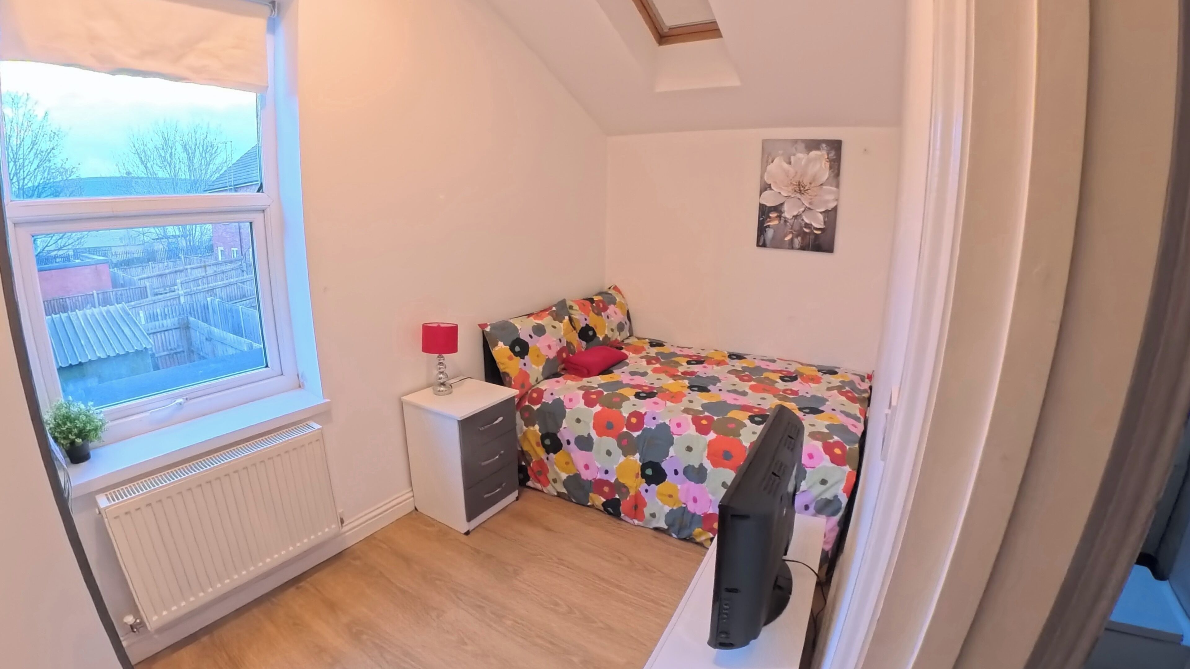A small bedroom with a single bed, a nightstand, a desk, a chair, a radiator, and a window with a view of buildings.