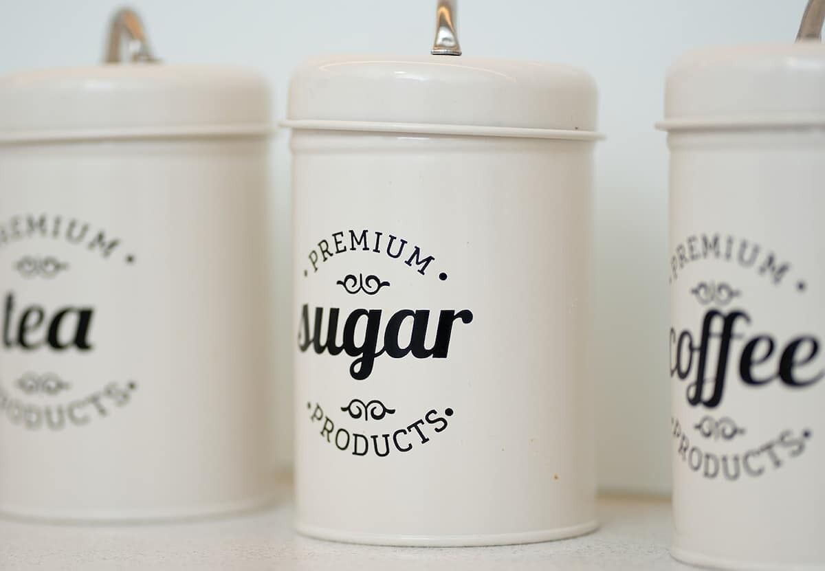 Tre vita behållare med lock, var och en märkt med namnet på en annan produkt: ”Te av högsta kvalitet”, ”Socker av högsta kvalitet” och ”Kaffe av högsta kvalitet”.