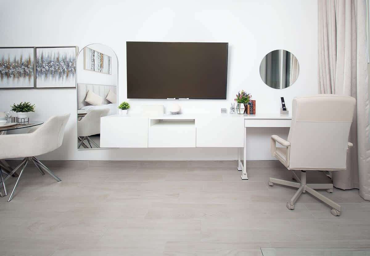 Una habitación moderna y minimalista con un escritorio blanco, un televisor de pantalla plana grande, un espejo redondo y una silla de oficina blanca.
