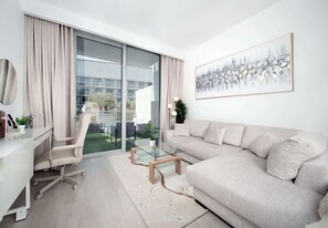 A modern living room with a glass coffee table, a beige sofa, a desk with a chair, and a large window with curtains.