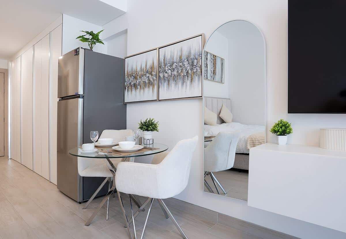 Een moderne keuken met een inbouwkoelkast, een eethoek met een glazen tafel en witte stoelen, en een zithoek met een bank en een grote spiegel.