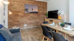 A modern living room with a brick wall, a flat-screen TV, a dining table set with plates and cups, and a sofa with a book and glasses on it. - Stunning views over iconic Sagrada Familia! Building perfect for large groups! (Barcelona)