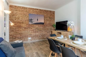 Ein modernes Wohnzimmer mit Backsteinwand, Flachbildfernseher, Esstisch gedeckt mit Tellern und Tassen sowie einer Couch mit Buch und Brille darauf.