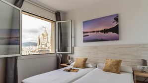A hotel room with a bed, a nightstand, a lamp, a window with a view of a cityscape and a building, and a painting on the wall. - Stunning views over iconic Sagrada Familia! Building perfect for large groups! (Barcelona)