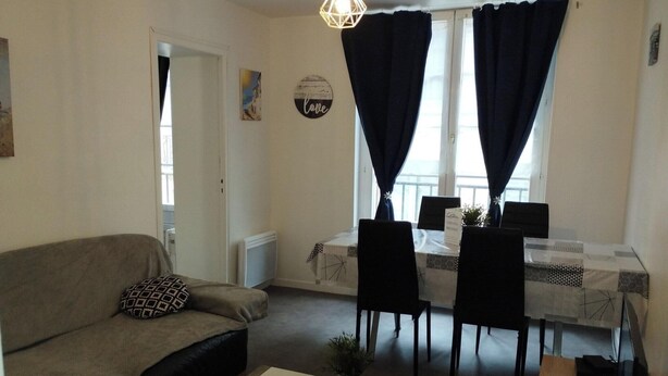 En stue med sofa, sofabord og spiseplads med bord og stole. Der er et vindue med gardiner, en radiator og en vægmonteret lampe.