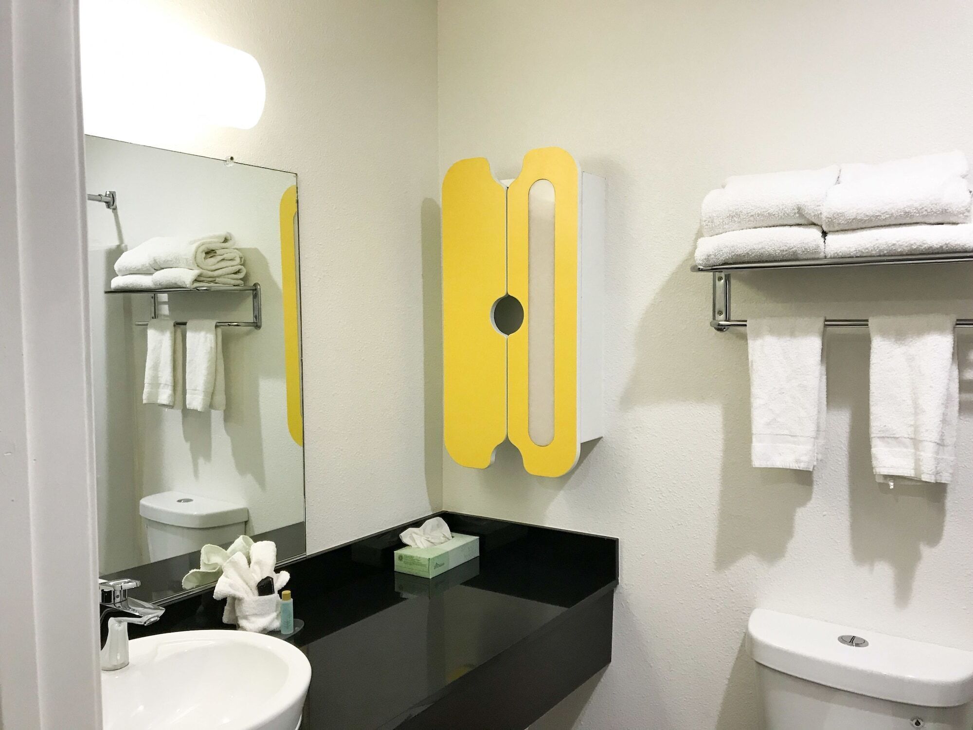 A modern bathroom with a black countertop, white sink, and a toilet. There are towels on a rack and a mirror with a yellow accent.