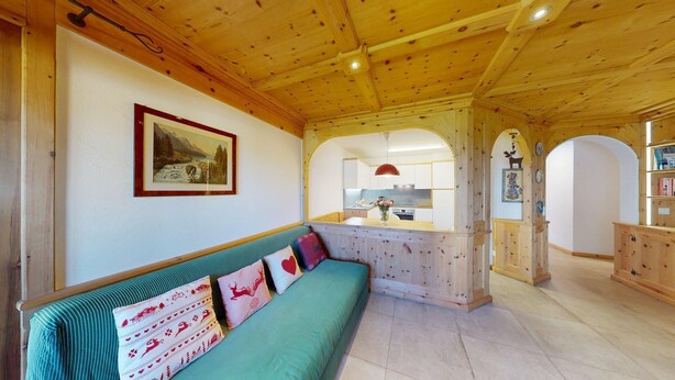 A cozy room with a wooden ceiling, a green sofa with patterned cushions, a framed picture on the wall, and a kitchen visible through an archway. - Centrally located residential complex in St. (St. Moritz)