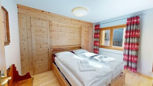 A bedroom with a wooden headboard, a bed with white linens, a window with red curtains, and a view of a building outside. - Centrally located residential complex in St. (St. Moritz)