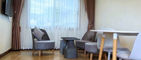 A modern living room with a TV mounted on the wall, two grey armchairs, a small round table, and a dining table with chairs.