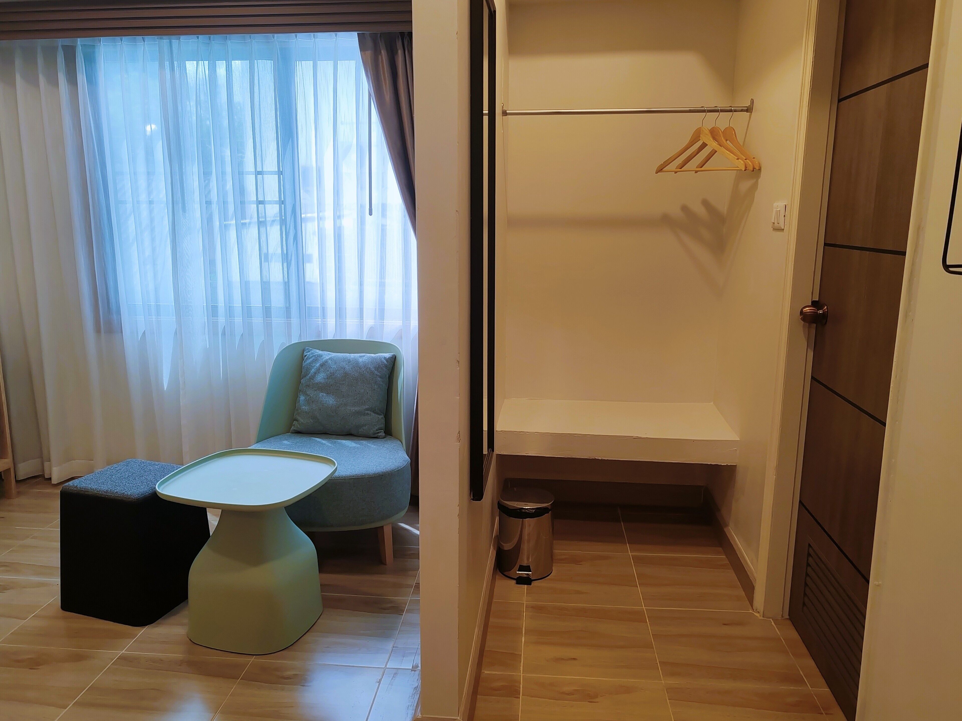 A room with a chair, a small table, a hanging clothes rack, and a trash bin.