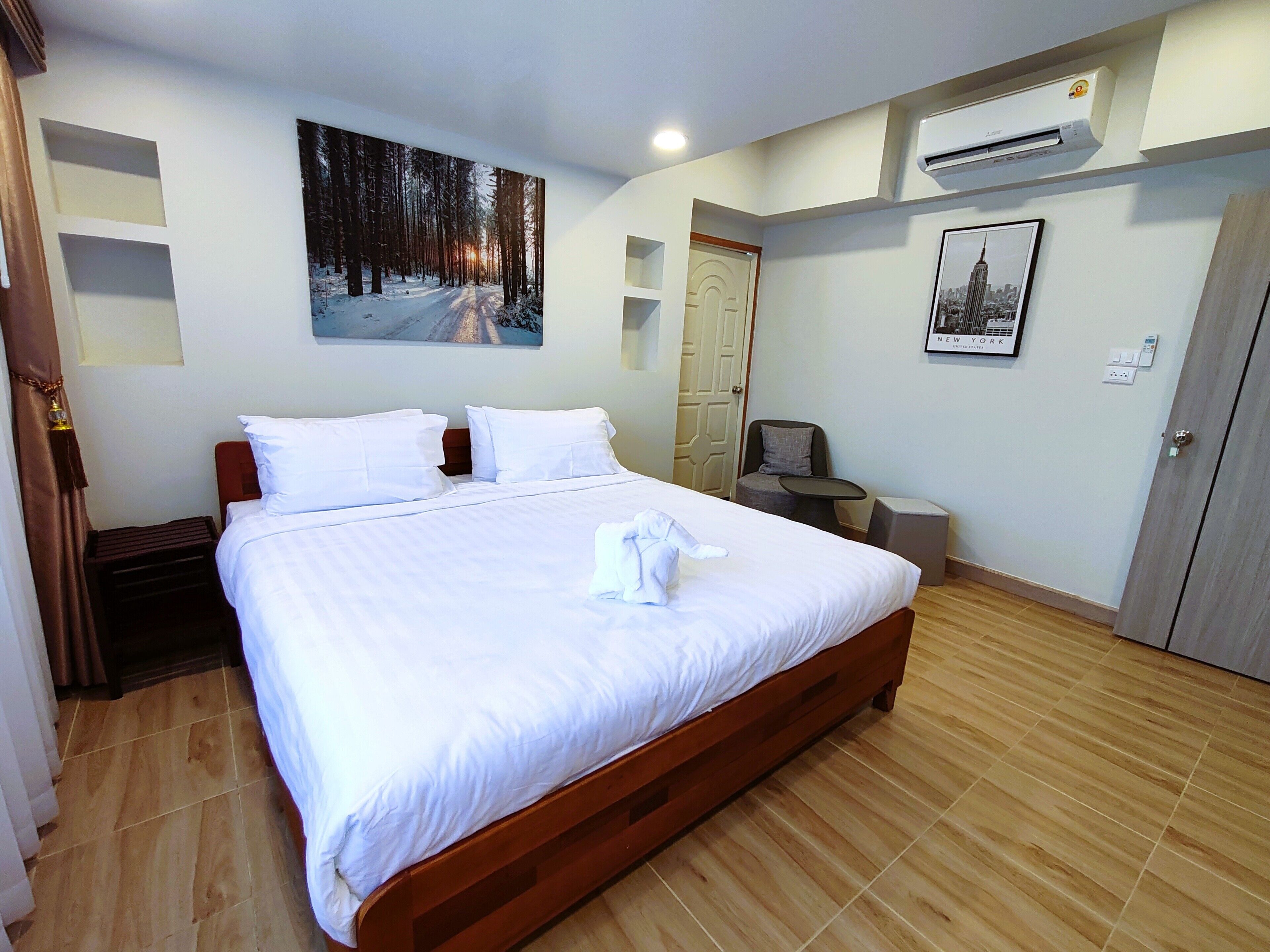 Habitación de hotel con una cama grande, un cuadro de un bosque nevado, un equipo de aire acondicionado, un retrato enmarcado de Nueva York y piso de madera.