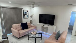 A modern living room with a sofa, a coffee table, a TV, and a ceiling fan. - Classy Comfy 3-bedroom house with AC in Palm Bay (Palm Bay)
