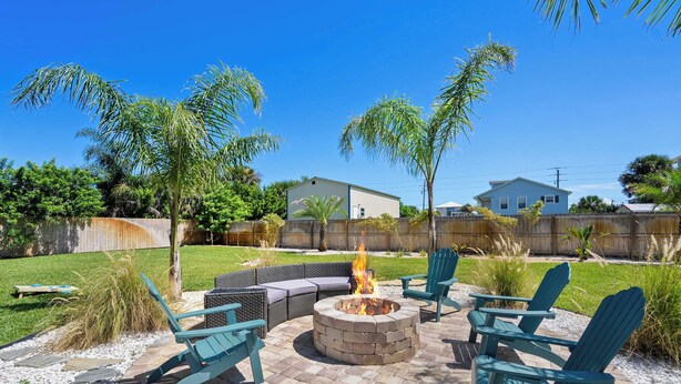 A patio with a fire pit, green Adirondack chairs, and a grassy yard with palm trees.