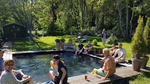 Un grupo de personas descansando junto a una piscina con cubierta de madera, rodeado de vegetación y un pequeño cobertizo.