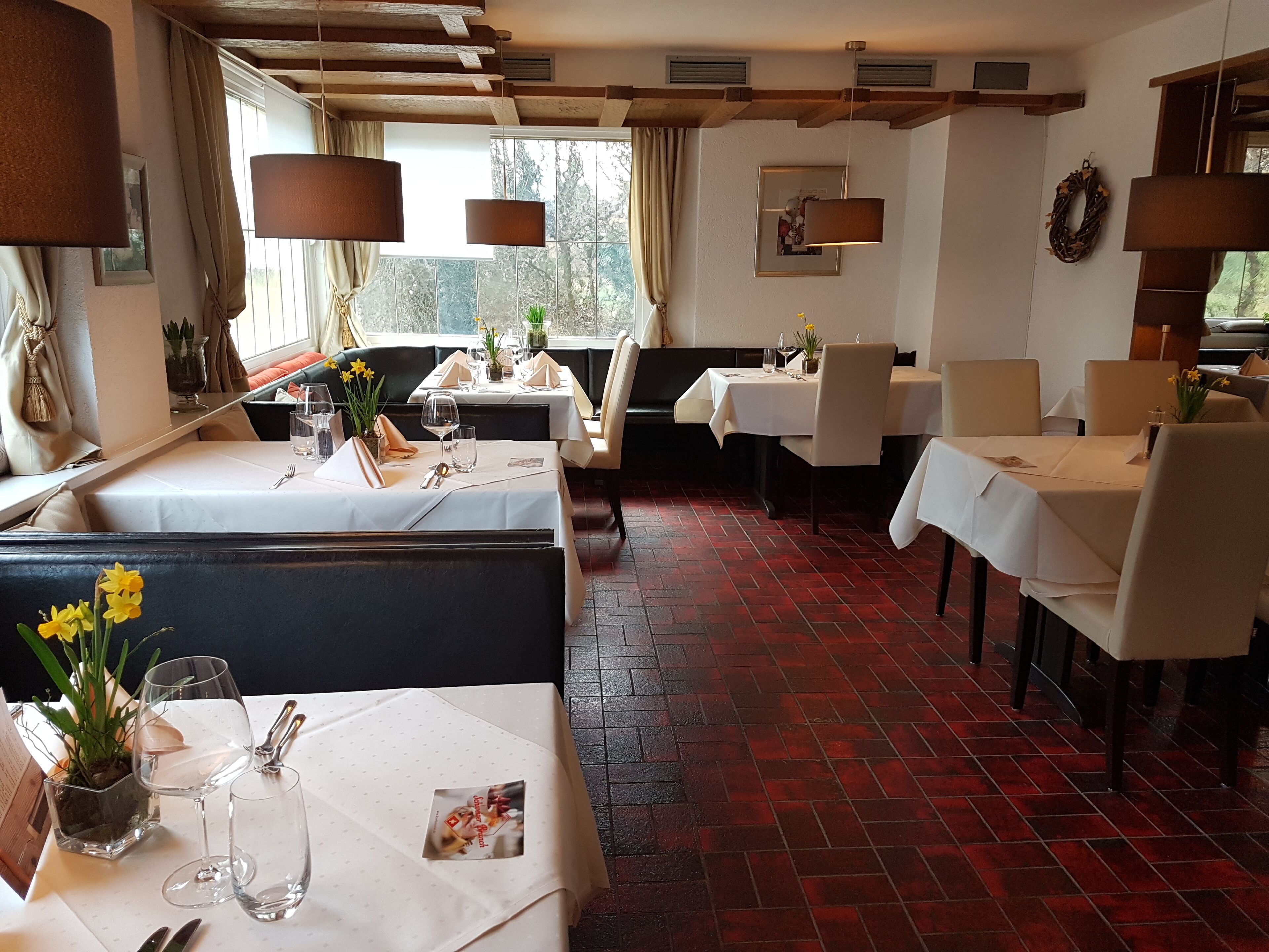 A restaurant with booths and tables, red brick flooring, and natural light from large windows.