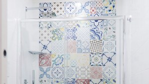 Zona de ducha con mampara de vidrio, un taburete de madera y una bañera blanca.