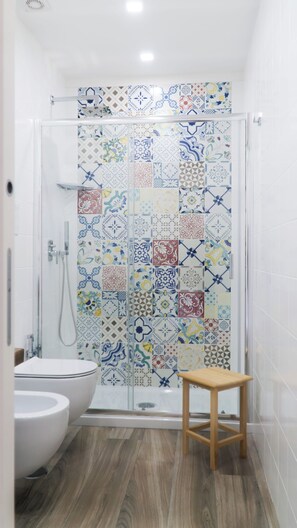 Zona de ducha con mampara de vidrio, un taburete de madera y una bañera blanca.