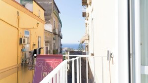 Una estrecha calle urbana con edificios a ambos lados, vista despejada al mar y un balcón con toldo morado.