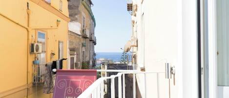 A narrow urban street with buildings on either side, a clear view of the sea, and a balcony with a purple awning.