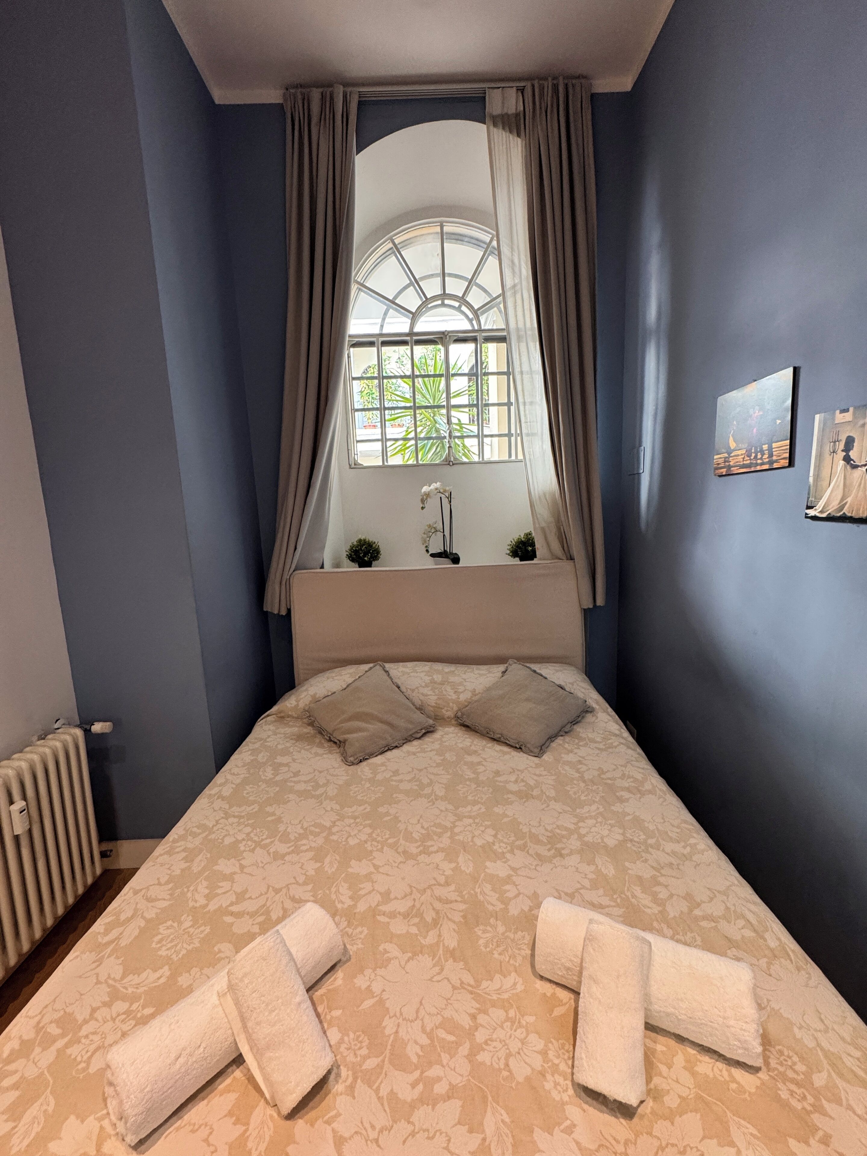 Een slaapkamer met een groot bed, een raam met gordijnen, een radiator en twee wanddecoraties.