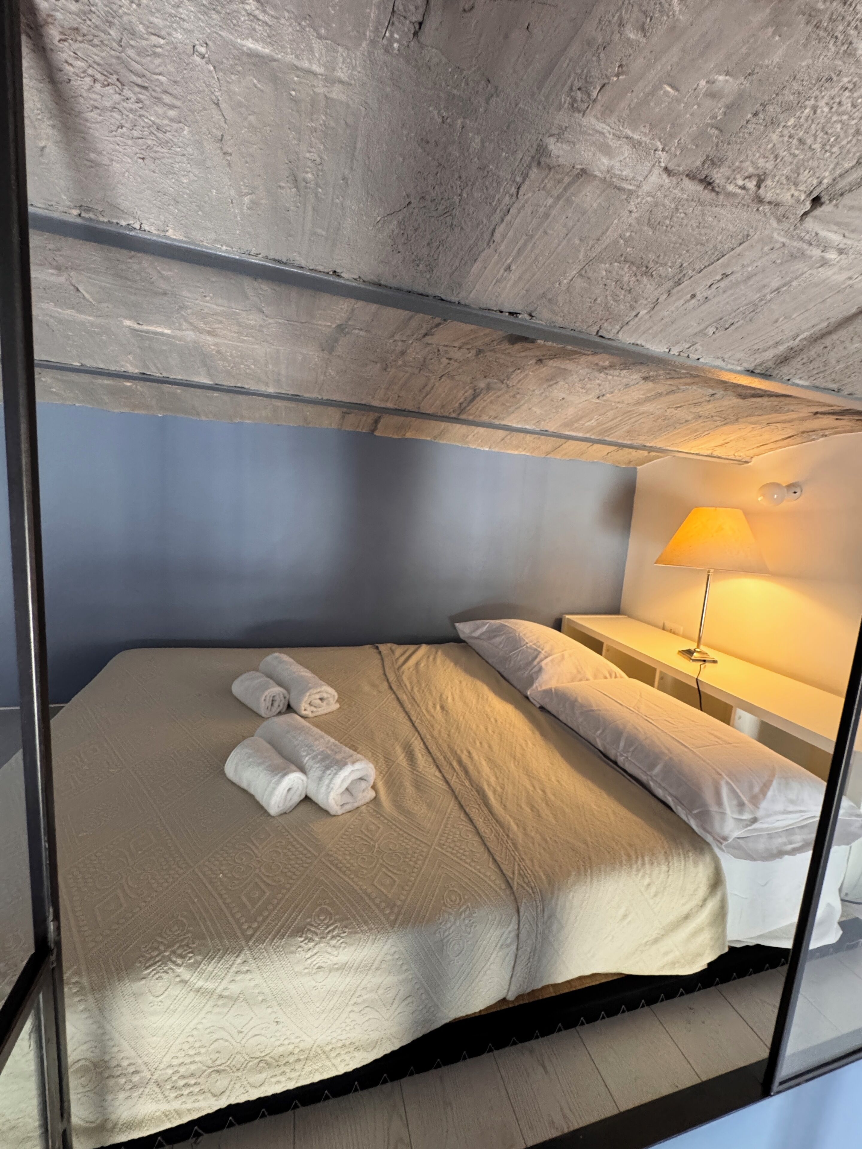 A single bed with rolled towels, a bedside table with a lamp, and a bunk bed above.