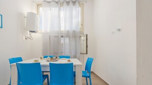 Una zona de menjador amb taula blanca i cadires blaves, una finestra amb cortines translúcides, un llum penjant del sostre i un quadre al mur.