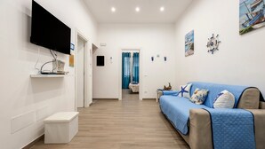 Habitació d'hotel moderna amb televisió de pantalla plana, un sofà amb funda blava, una petita taula de nit i una porta que dona a una altra habitació.