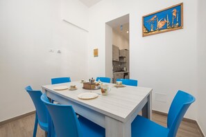 Un coin repas avec une table dressée pour quatre personnes, des chaises bleues, une cuisine visible à travers une porte ouverte et une œuvre d’art encadrée accrochée au mur.