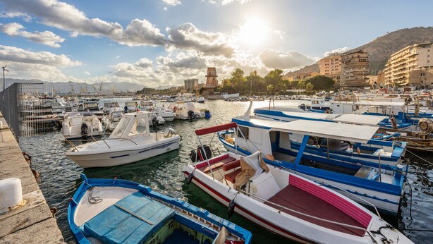 Un port esportiu amb diverses embarcacions amarrades, cel blau i edificis al fons.