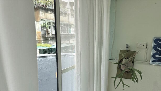Ein Zimmer mit Blick auf eine Straße, ein Fenster mit weißen Vorhängen, eine Wandsteckdose und ein Blumentopf.