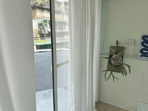 Een kamer met uitzicht op een straat, een raam met witte gordijnen, een wandcontactdoos en een kamerplant.