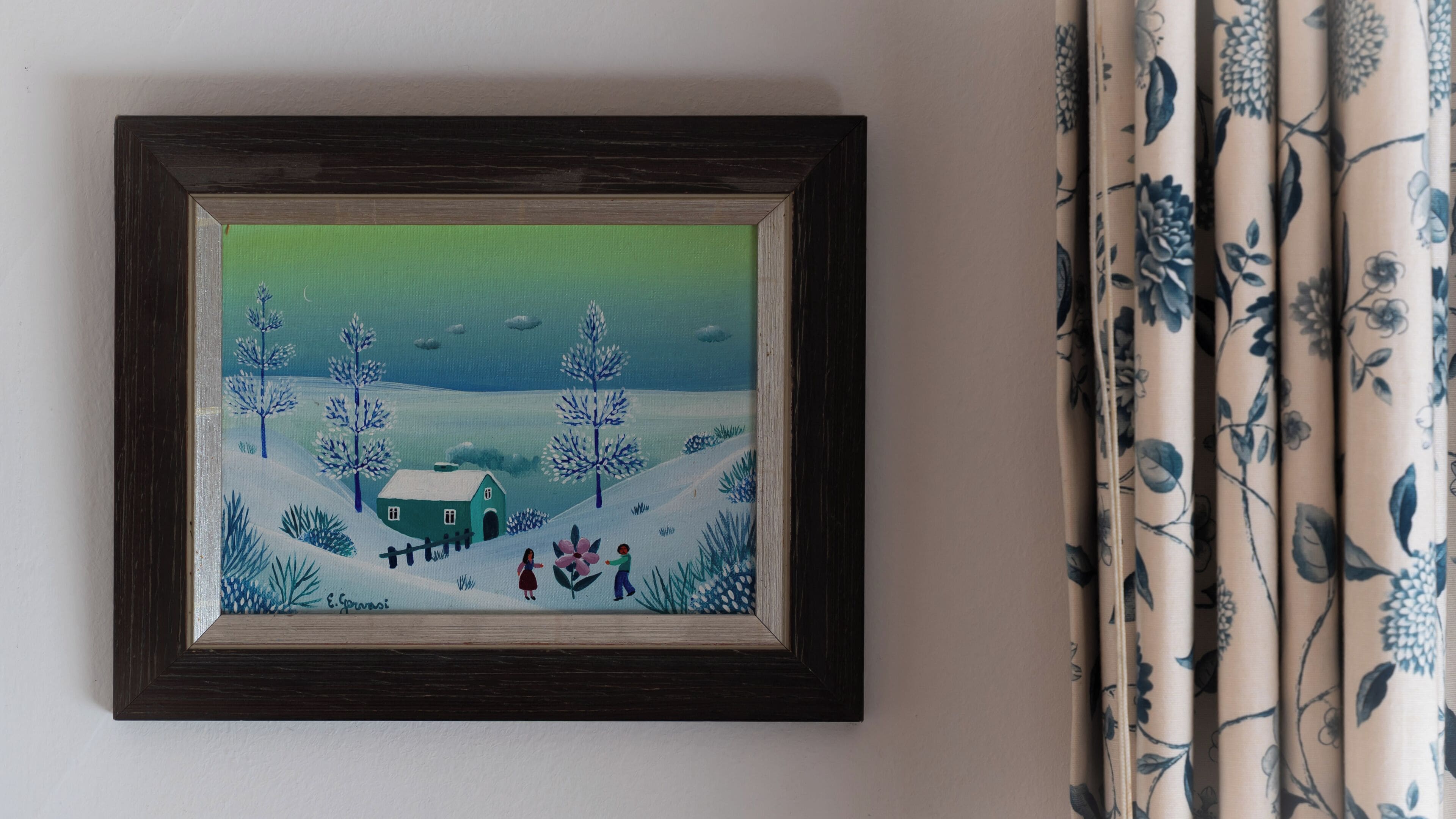 Un tableau encadré représentant un paysage enneigé avec une maison et deux personnages, accroché au mur à côté d’un rideau orné de motifs floraux.