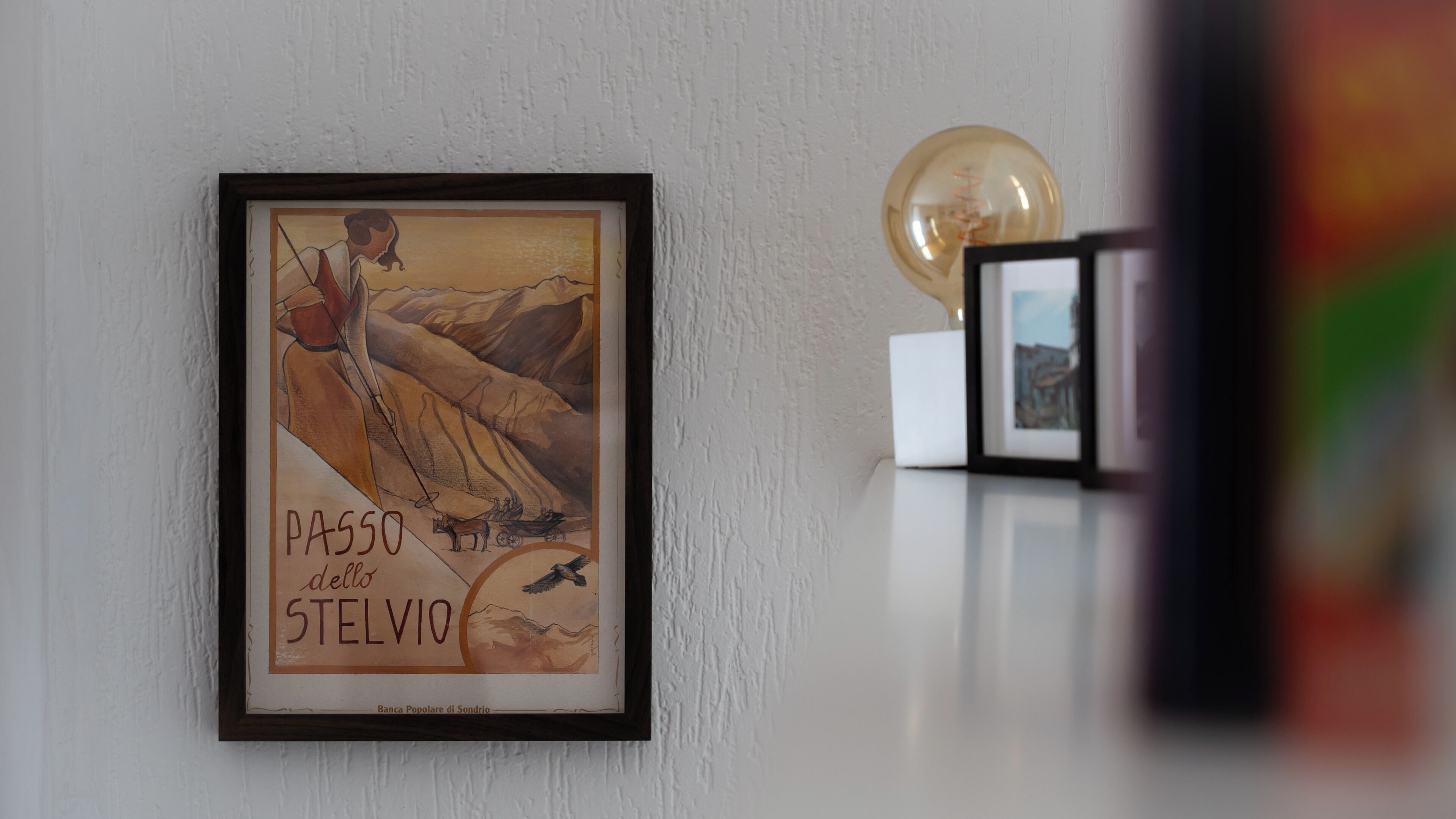 A framed artwork titled 'Passo del Stelvio' on a white wall, with a light bulb and a framed photo in the background.