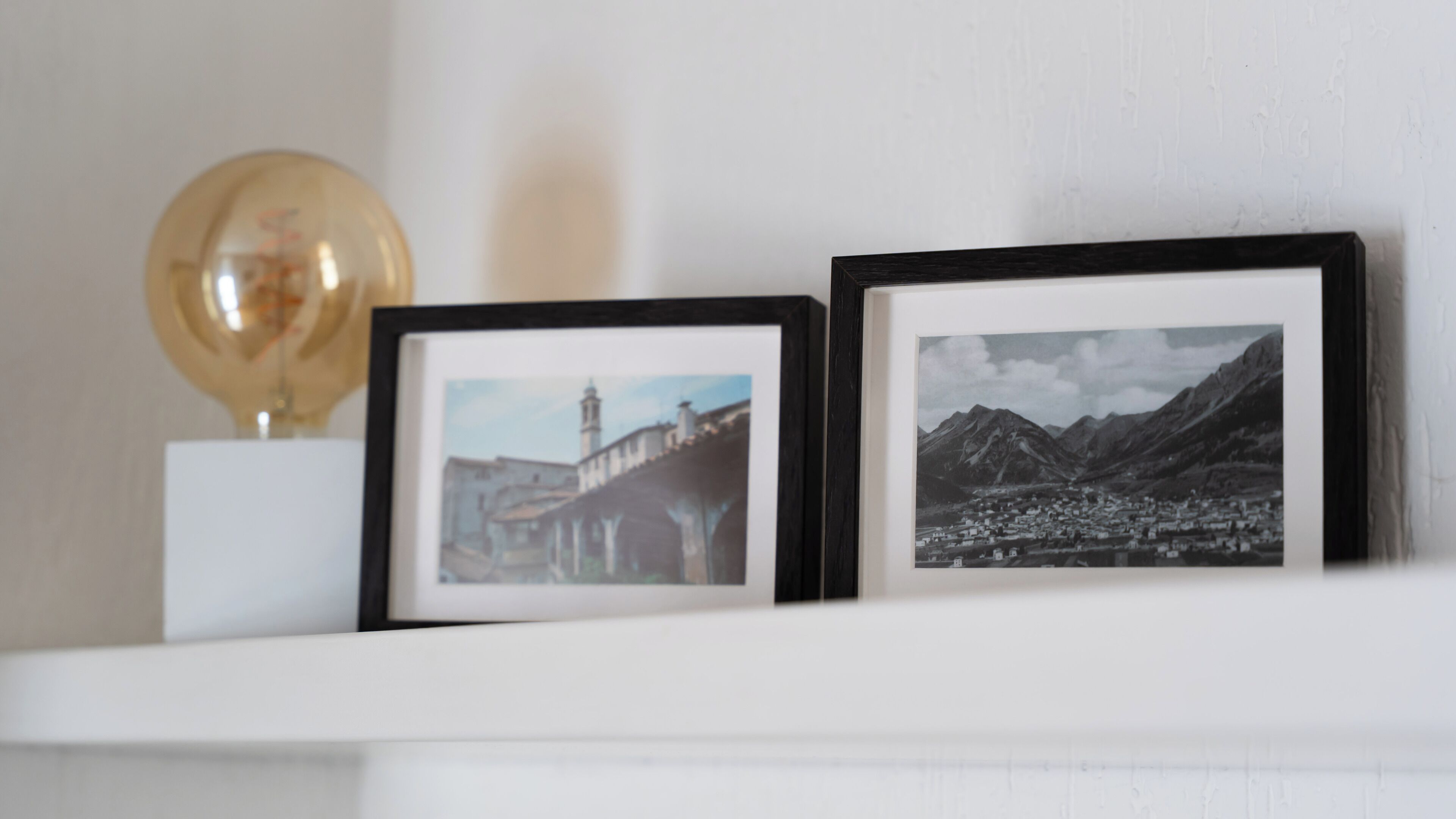 Two framed photographs on a white shelf, one depicting a bridge and the other a mountainous landscape.