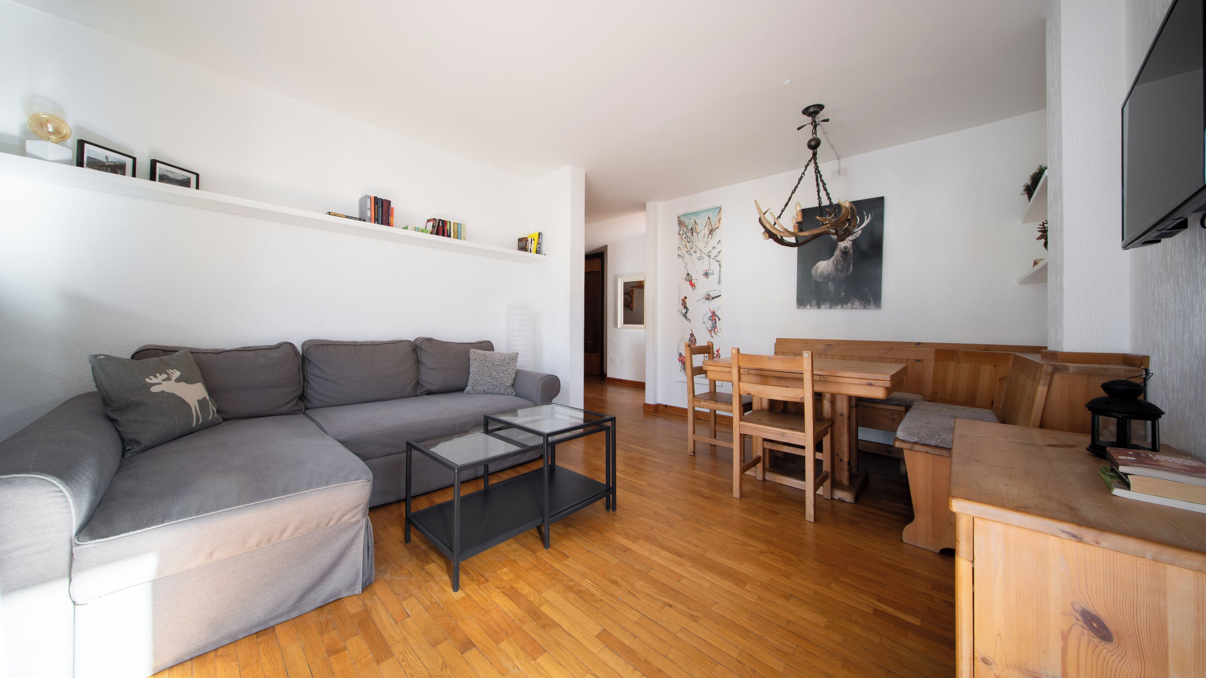 A modern living room with a grey sectional sofa, a glass coffee table, a wooden dining table with chairs, and a chandelier.