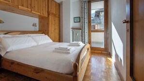 A wooden bed with white linens and towels, a wooden wardrobe, a radiator, and a view of a building through a glass door.