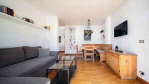 A modern living room with a grey sofa, a glass coffee table, wooden flooring, a wooden dining table with benches, and a mounted TV.