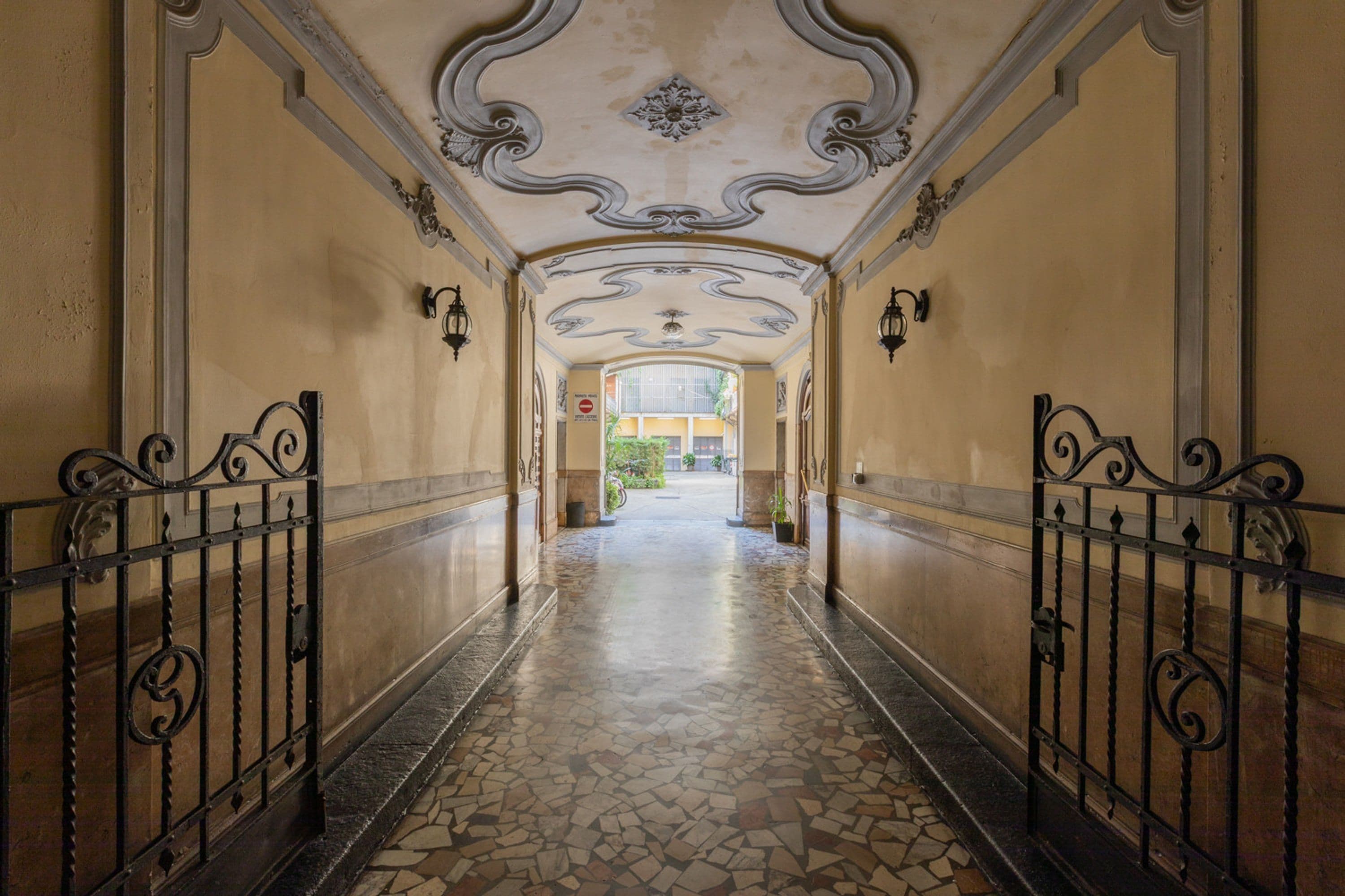 A corridor with ornate ceiling and tiled flooring, leading to a doorway with a view of greenery outside.