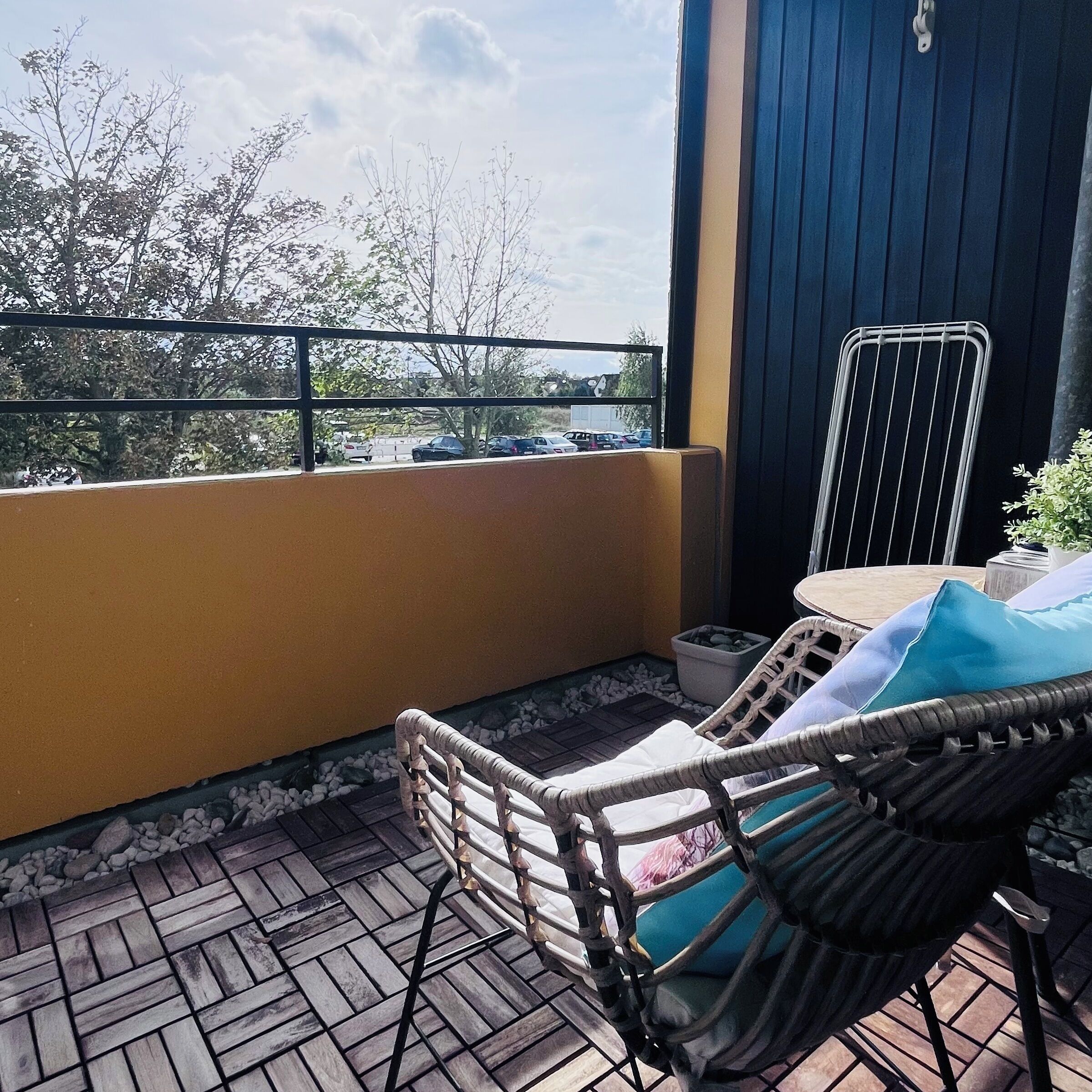Ein Balkon mit einem Korbstuhl und einem Tisch, Blick auf den Parkplatz und ein Gebäude mit blauer Fassade.