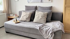 A modern living room with a grey sofa, two grey cushions, a wooden side table with a wine glass and bottle, and a framed picture on the wall. - Cozy Apartment with Stunning Views in Marina Wendtorf (Wendtorf)