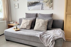 Uma sala de estar moderna com um sofá cinzento, duas almofadas cinzas, uma mesa lateral de madeira com uma taça e garrafa de vinho, e um quadro na parede.