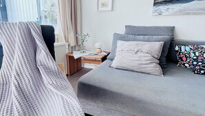 A modern bedroom with a grey sofa, a bedside table with a lamp, and a window with curtains. - Cozy Apartment with Stunning Views in Marina Wendtorf (Wendtorf)