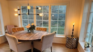 Un coin repas comprenant une table ronde en bois, six chaises rembourrées, un lustre et une fenêtre donnant sur la forêt.