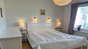 Une chambre à coucher bien rangée, avec un grand lit, deux tables de chevet avec des lampes, une table de nuit en bois, une commode blanche et une fenêtre avec des rideaux.