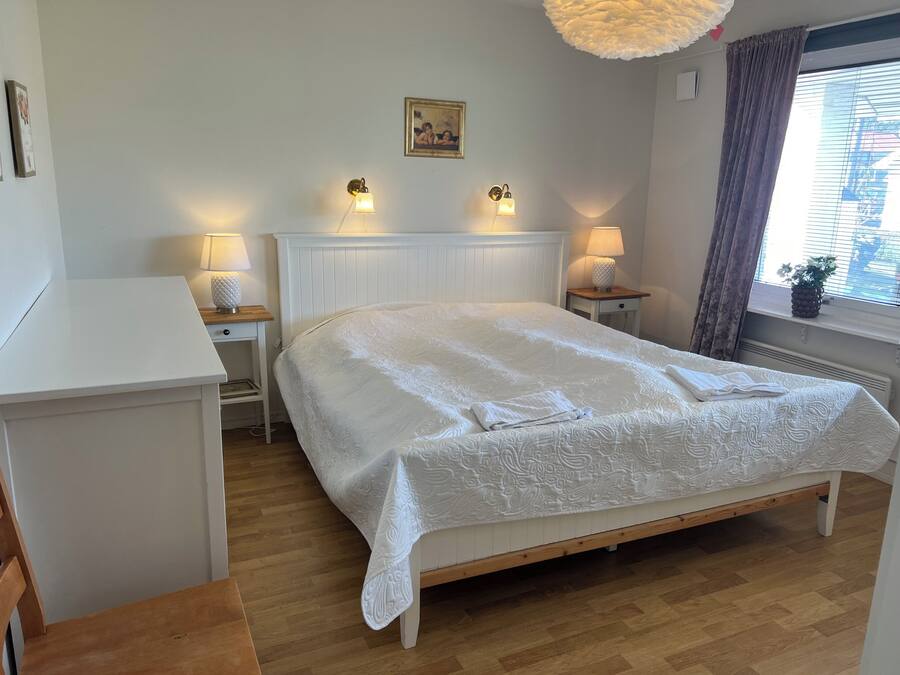 A neatly arranged bedroom with a large bed, two bedside tables with lamps, a wooden nightstand, a white dresser, and a window with curtains.