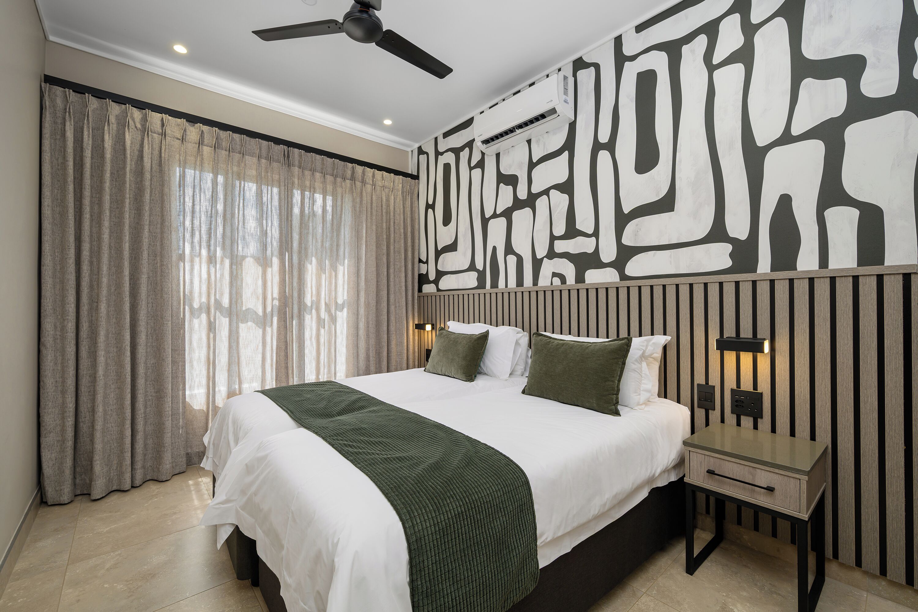 Ein Schlafzimmer mit einem Bett, Vorhängen, einem Deckenventilator, einem Nachttisch und einer Wand mit geometrischem Muster.