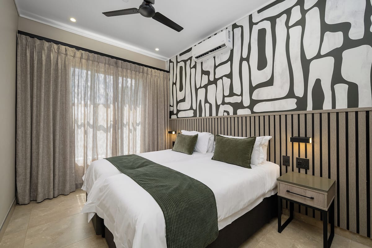 A bedroom with a bed, curtains, a ceiling fan, a bedside table, and a wall with a geometric pattern.