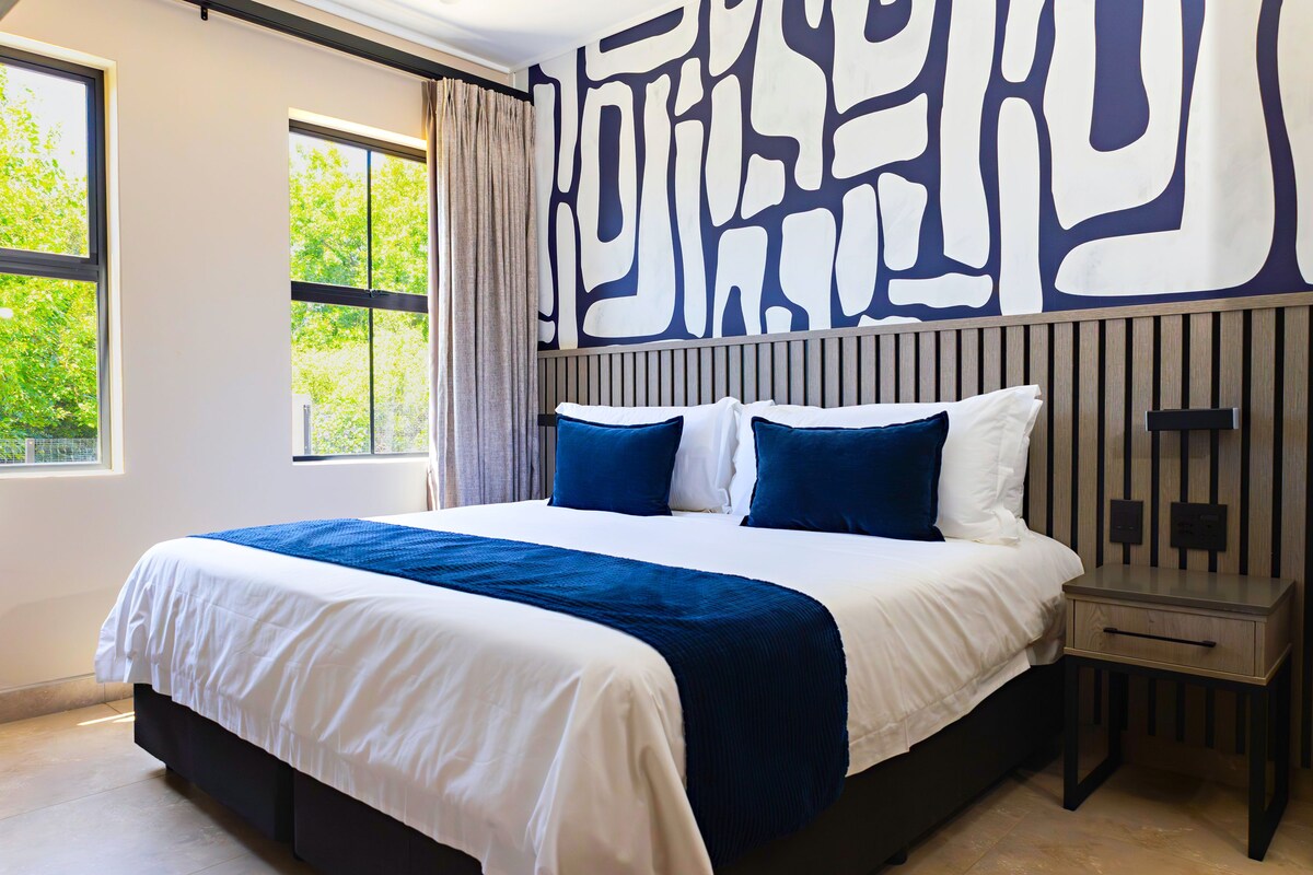 A modern hotel room with a large bed, a nightstand, and a wall with abstract art.