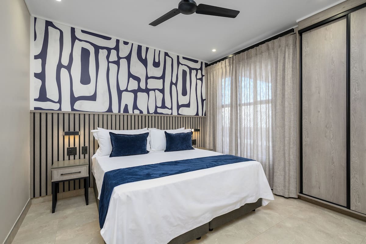 A modern bedroom with a large bed, a ceiling fan, a nightstand, and a wall with abstract art.